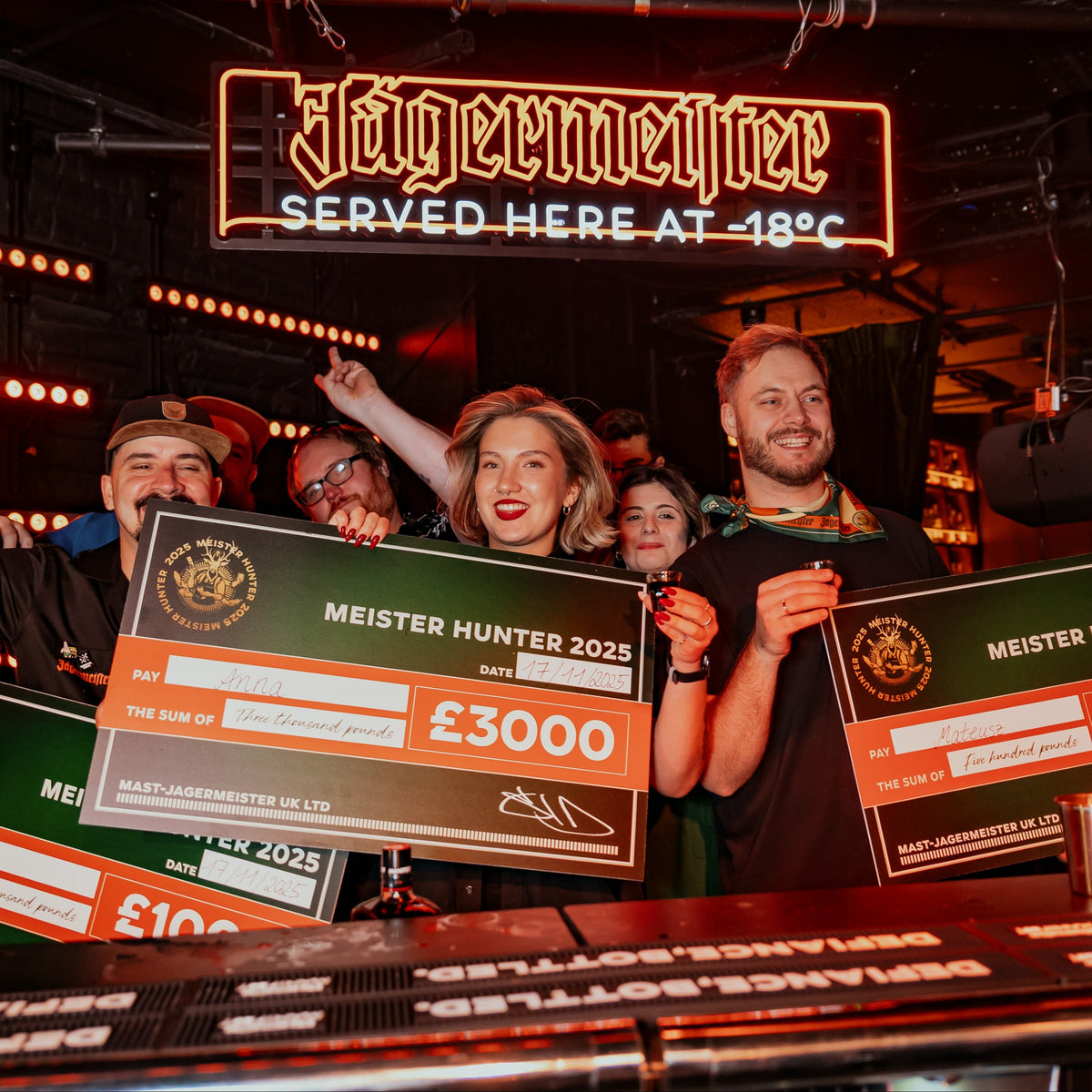 A group of people celebrating in a bar or event space, each holding a large novelty check from Meister Hunter 2025 for £3000. The background features a glowing 'Jägermeister' sign above them.