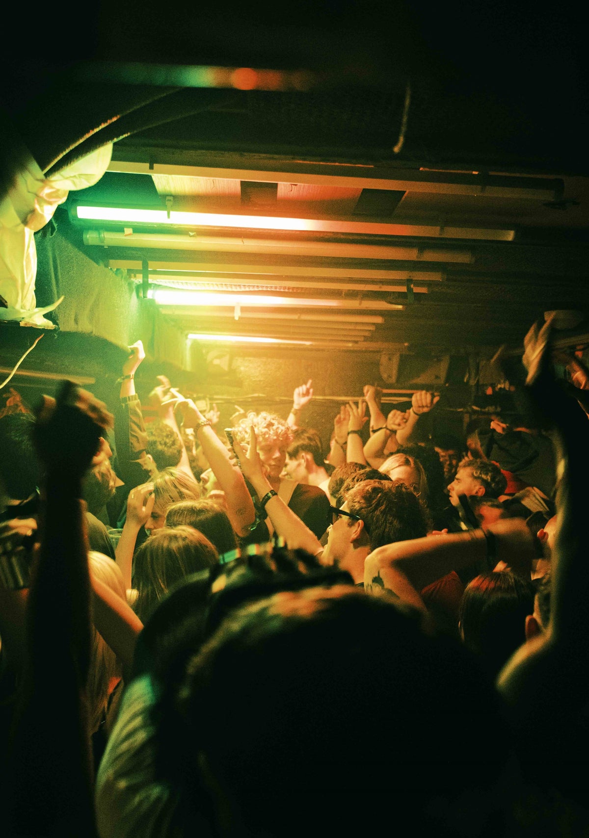 A crowded nightclub or concert venue with a group of people dancing and raising their hands under green and orange stage lighting.