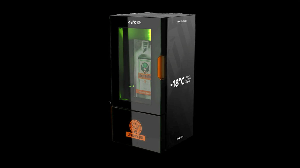 A black glass-door temperature-controlled storage cabinet with a green interior light and an orange accent, shown on a solid black background. The cabinet features white text reading '-18°C' and an orange Jägermeister logo near the bottom.