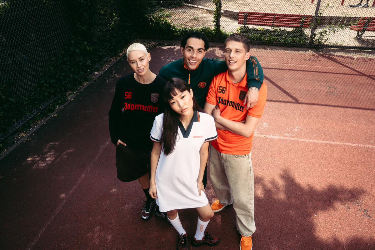 Four people posing together on an outdoor red tennis court, wearing matching dark green and orange Jägermeister athletic jerseys. The group includes a person with platinum blonde hair in a black jersey, a man in an orange jersey, a woman in a white polo dress, and another man in a dark green jersey.