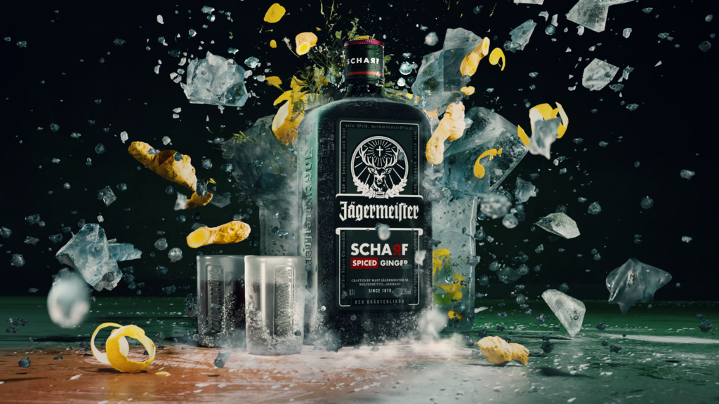 A black bottle of Jägermeister, labeled "SCHARF", is surrounded by two shot glasses, ice, and lemon peels suspended in the air. Water splashes around the items on a dark surface.