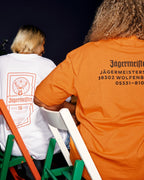 Twee mensen zitten met hun rug naar de camera op groene en witte stoelen, met Jägermeister-T-shirts aan. Het ene shirt is wit met een oranje labelprint, en het andere is feloranje met het adres van het hoofdkantoor van Jägermeister in zwart.