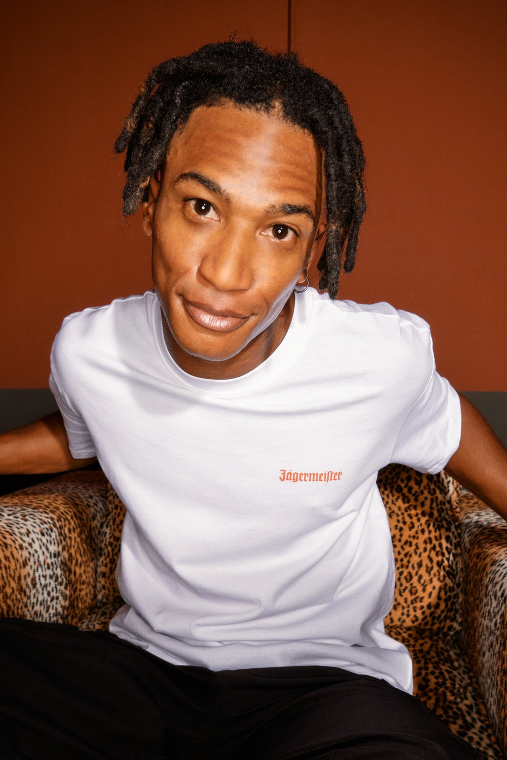 Persoon in een wit T-shirt met een Jägermeister-logo op de borst, zittend op een stoel met patroon tegen een oranje muur.