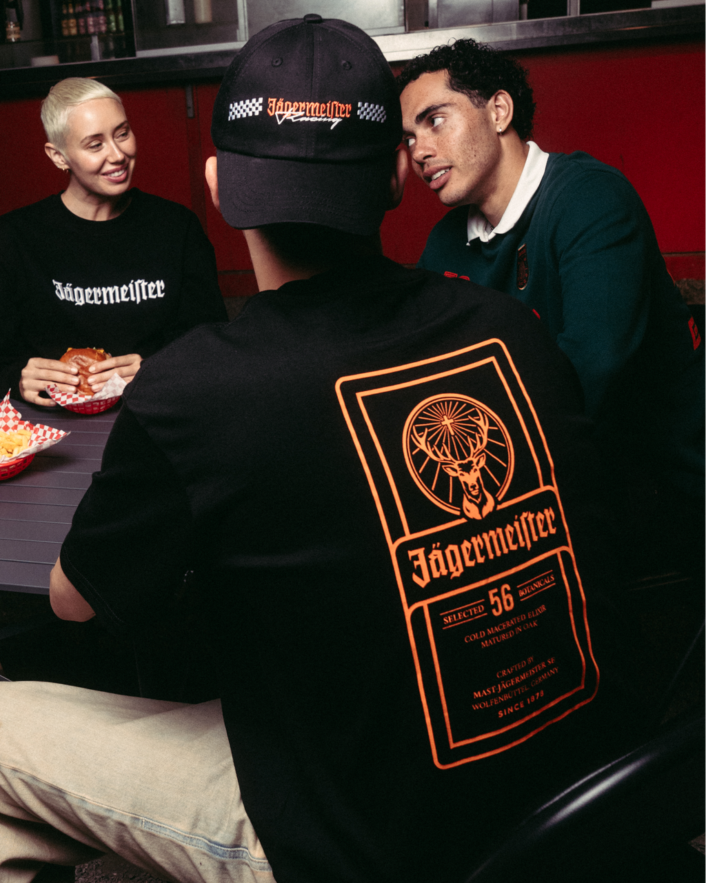 Vier mensen verzamelen zich rond een tafel bij een eetkraam, met de focus op een persoon die een zwart Jägermeister-T-shirt draagt met een groot oranje graphic op de achterkant, kaki broek en een zwarte pet. 