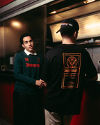 Twee mannen staan 's nachts buiten bij een rode foodtruck. De ene man kijkt naar de camera en draagt een donkergroene Jägermeister Longsleeve en een zwarte broek. De andere draagt de Jägermeister Racing Cap en het zwarte Jägermeister-label T-shirt.