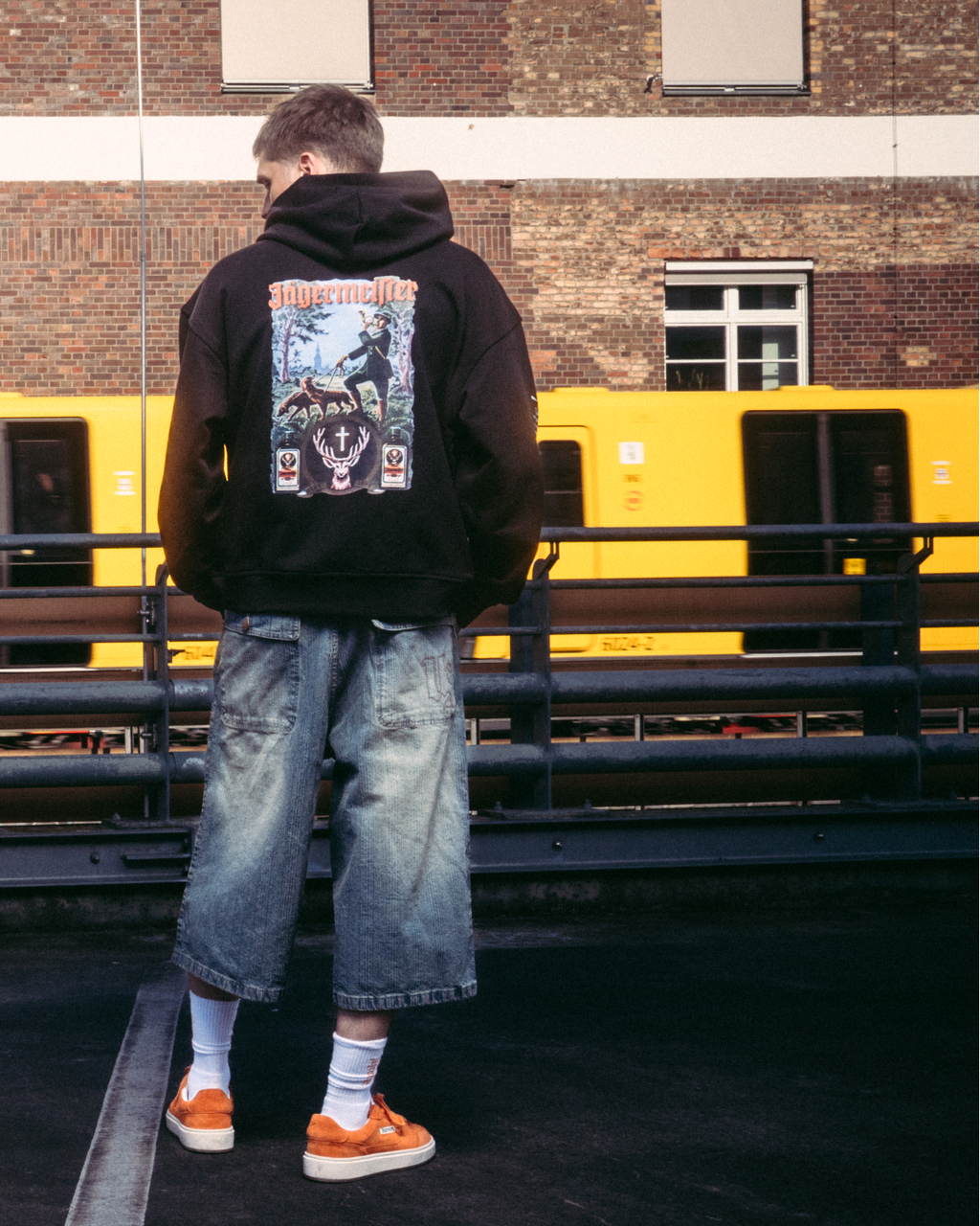 Een model in streetwear, waaronder een zwarte hoodie met de Jägermeister tradition-afbeelding op de achterkant, staat op een donker verharde ondergrond voor een bakstenen gebouw en een gele trein.