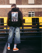 Een model in streetwear, waaronder een zwarte hoodie met de Jägermeister tradition-afbeelding op de achterkant, staat op een donker verharde ondergrond voor een bakstenen gebouw en een gele trein.