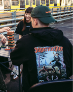Vier mensen zitten buiten rond een klaptafel en spelen een stapelspel. Op de achtergrond is achter een metalen railing een gele en witte trein te zien. De persoon op de voorgrond draagt de Jägermeister Tradition Hoodie en een groene Jägermeister Dad Cap.