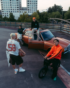Een groep van vier mensen in een stedelijke buitenomgeving met een roestige oranje auto, moderne hoogbouw en een brug op de achtergrond. Eén persoon in een feloranje sweatshirt zit op een fiets op de voorgrond, een ander staat in een wit Jägermeister-jersey met '56' erop en houdt een voetbal vast, en twee anderen staan rond de vintage auto.