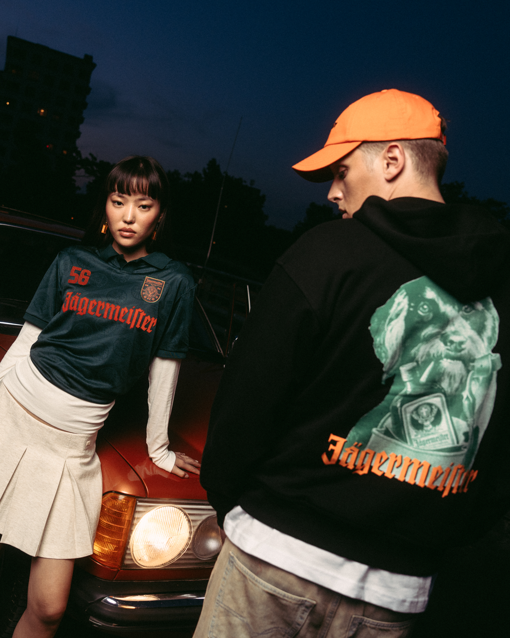 Twee modellen poseren voor een vintage rode auto in de schemering. Eén model draagt een donkergroen Jägermeister-shirt met een witte geplooide rok, en de ander draagt een zwarte Jägermeister Waldi hoodie en een oranje Jägermeister Dad cap.