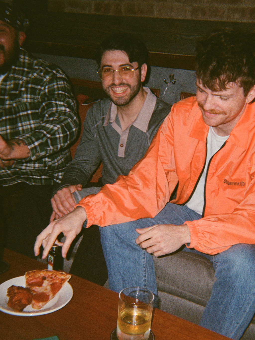 Twee mannen die samen zitten, één draagt een oranje Jägermeister-jas, met een pizzabord en een glas op tafel.