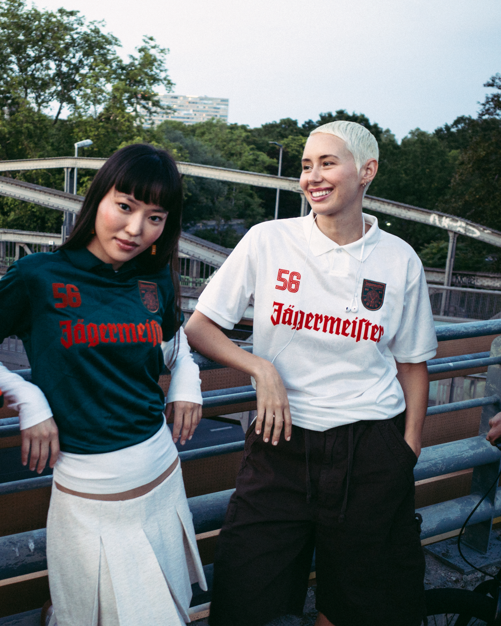 Vier mensen staan buiten op een brug of wandelpad en dragen streetwear. Eén persoon draagt een feloranje zip-up jas, een ander draagt een donkergroen Jägermeister-shirt met een witte rok, een derde draagt een wit Jägermeister-shirt met donkere shorts, en een vierde draagt een zwarte hoodie en een oranje pet.