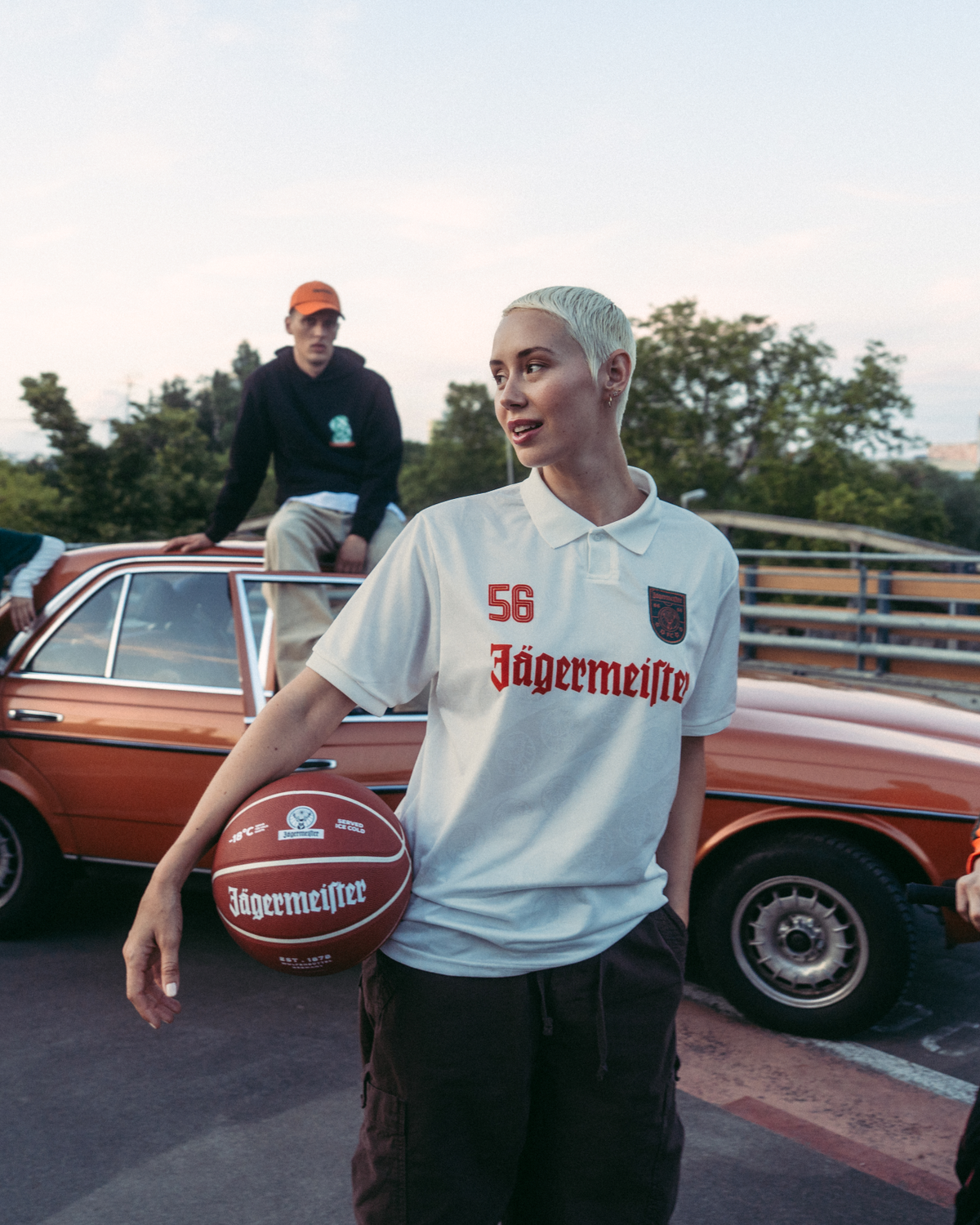 Een groep van vier mensen poseert buiten op een brug of viaduct, met achter hen een vintage oranje auto. De persoon in het midden houdt een basketbal vast en draagt een wit Jägermeister-shirt, terwijl de anderen feloranje, groene en zwarte streetwear dragen.