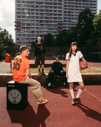Lifestyle-scène in de buitenlucht op een basketbalveld met een hoog appartementsgebouw op de achtergrond. Een zittende man in een oranje Jägermeister-shirt zit op een zwarte speakerachtige box, terwijl een staande vrouw in een witte Jägermeister polojurk een basketbal vasthoudt.