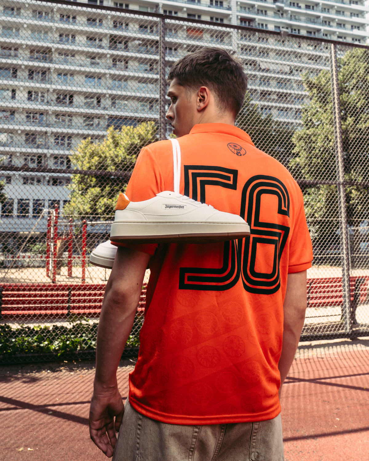 Een persoon staat op een buitensportveld met een feloranje Jägermeister-shirt met een grote “56” op de achterkant. Over één schouder hangt een paar witte Jägermeister-sneakers, met een gaashek en appartementsgebouwen op de achtergrond.