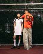 Twee modellen staan op een rode tennisbaan voor een gaashek. Het model links draagt een witte sportieve Jägermeister polojurk met donkergroene details, witte sokken en zwarte schoenen.