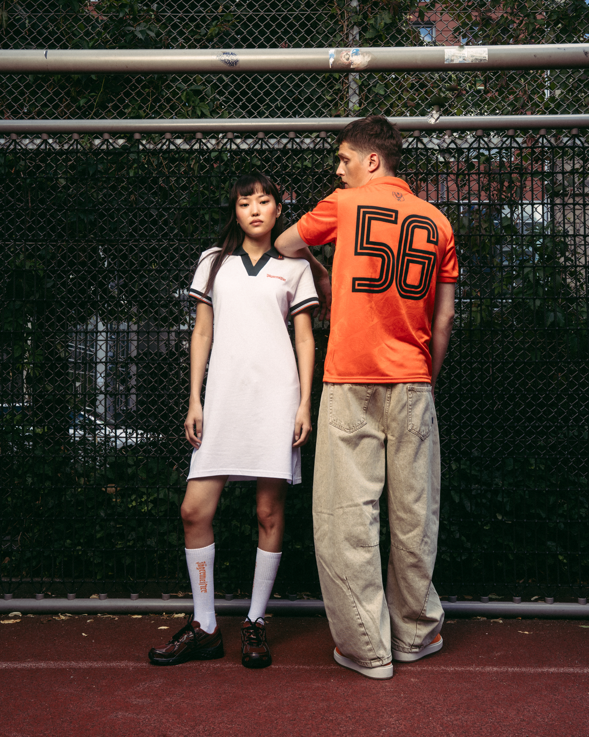 Twee modellen staan op een rode tennisbaan voor een gaashek. Het model links draagt een witte sportieve Jägermeister polojurk met donkergroene details, witte sokken en zwarte schoenen.