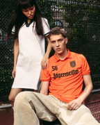 Een man in een feloranje Jägermeister-shirt en beige broek zit op een baan, terwijl een vrouw in een witte Jägermeister polojurk achter hem staat tegen een gaashek.