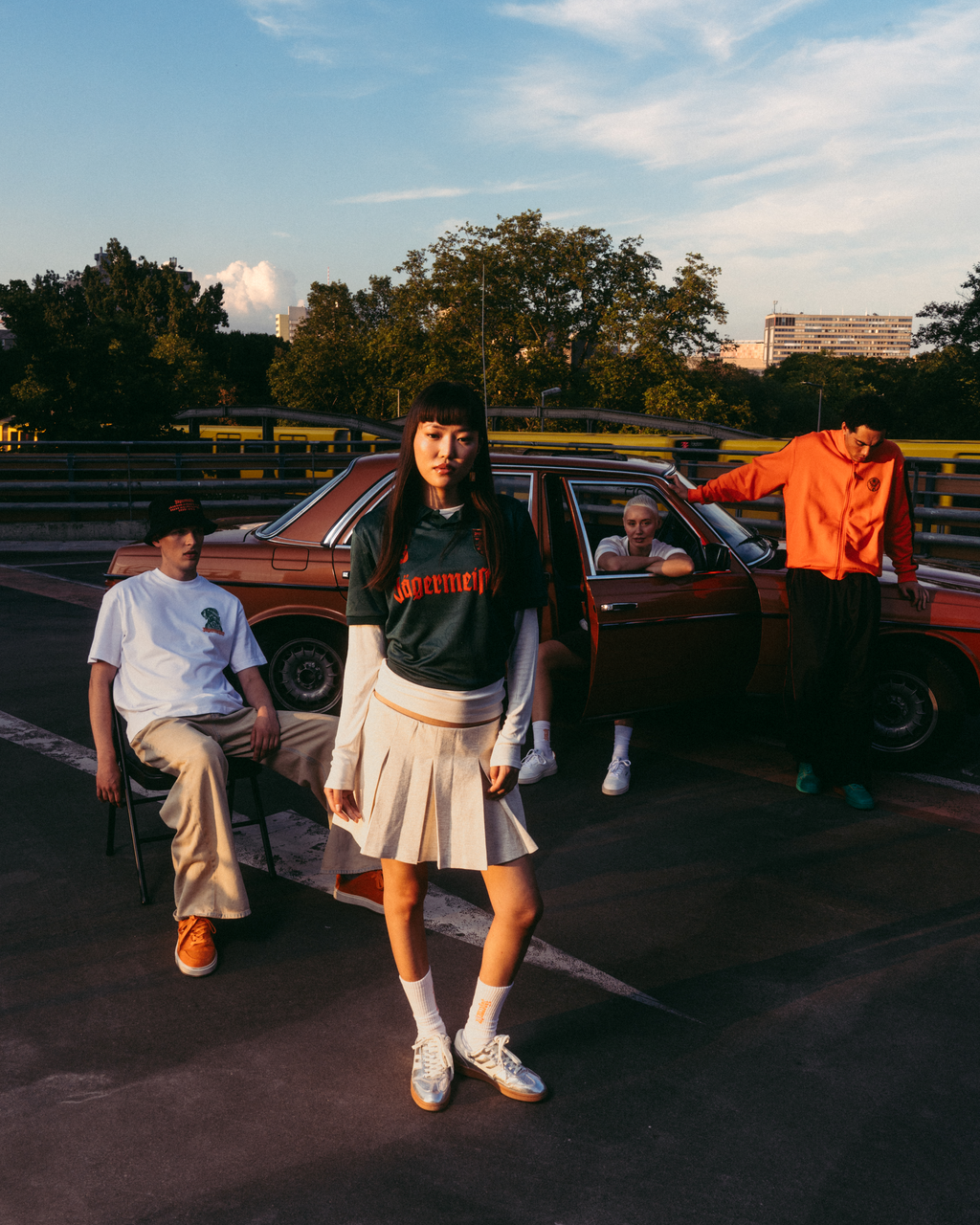 Een groep van vier jonge mensen poseert buiten op een racebaan bij zonsondergang naast een vintage rode auto. De persoon in het midden draagt een donkergroen Jäermeister-shirt met oranje letters, een geplooide witte rok en witte sneakers.