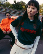 Een groep van vier mensen poseert buiten voor een vintage bruine auto. De persoon in het midden draagt een donkergroen Jägermeister-shirt, terwijl de anderen casual streetwear dragen.