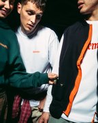 Drie jonge mannen staan dicht bij elkaar in een donkere setting en dragen Jägermeister-kleding. Eén draagt een donkergroene hoodie, een ander een wit shirt met lange mouwen en de derde een zwart-oranje jack met rits over een wit T-shirt.
