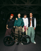 Vier jonge mannen poseren met een zwarte BMX-fiets in een donkere, industriële brugsetting bij nacht. Ze dragen Jägermeister-streetwear, waaronder een groene hoodie, wit T-shirt, zwarte bodywarmer en cargobroeken.