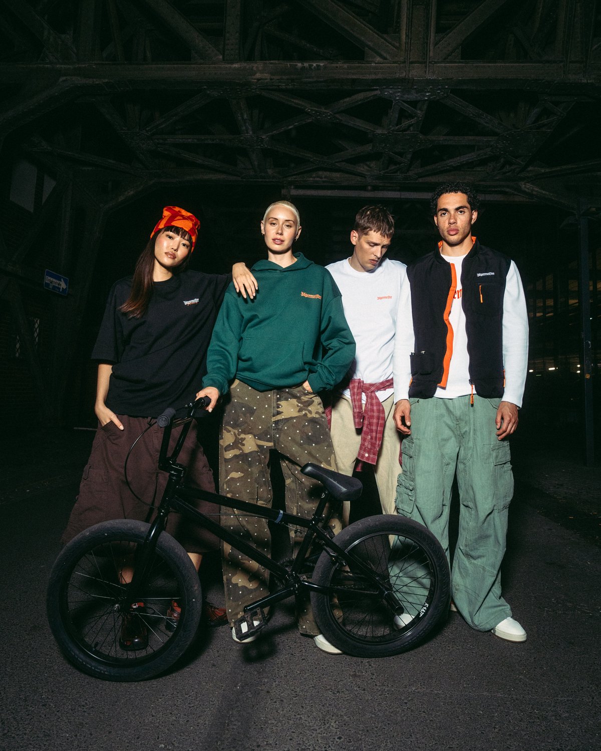 Vier jonge mannen poseren met een zwarte BMX-fiets in een donkere, industriële brugsetting bij nacht. Ze dragen Jägermeister-streetwear, waaronder een groene hoodie, wit T-shirt, zwarte bodywarmer en cargobroeken.