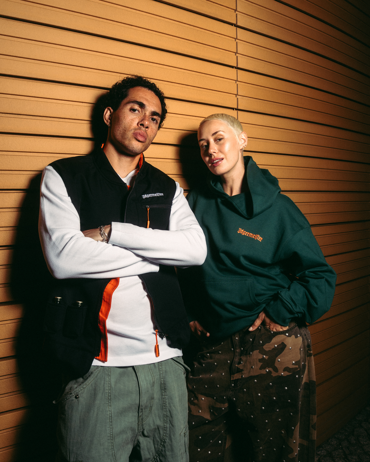 Twee modellen poseren tegen een warme wand met houten panelen. Het model links draagt een zwarte Jägermeister-bodywarmer over een wit shirt met lange mouwen en oranje accenten, en een olijfgroene cargobroek.