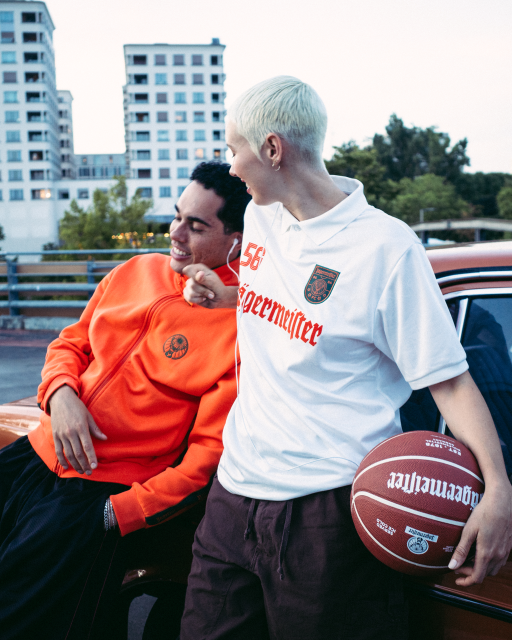 Twee mensen staan naast een vintage rode auto op een buitenparkeerplaats in de schemering. De man draagt een feloranje Jägermeister-sweatshirt en een zwarte broek, terwijl de vrouw een wit Jägermeister-shirt en een donkere broek draagt en een basketbal vasthoudt.