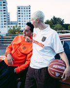 Twee mensen staan naast een vintage rode auto op een buitenparkeerplaats in de schemering. De man draagt een feloranje Jägermeister-sweatshirt en een zwarte broek, terwijl de vrouw een wit Jägermeister-shirt en een donkere broek draagt en een basketbal vasthoudt.