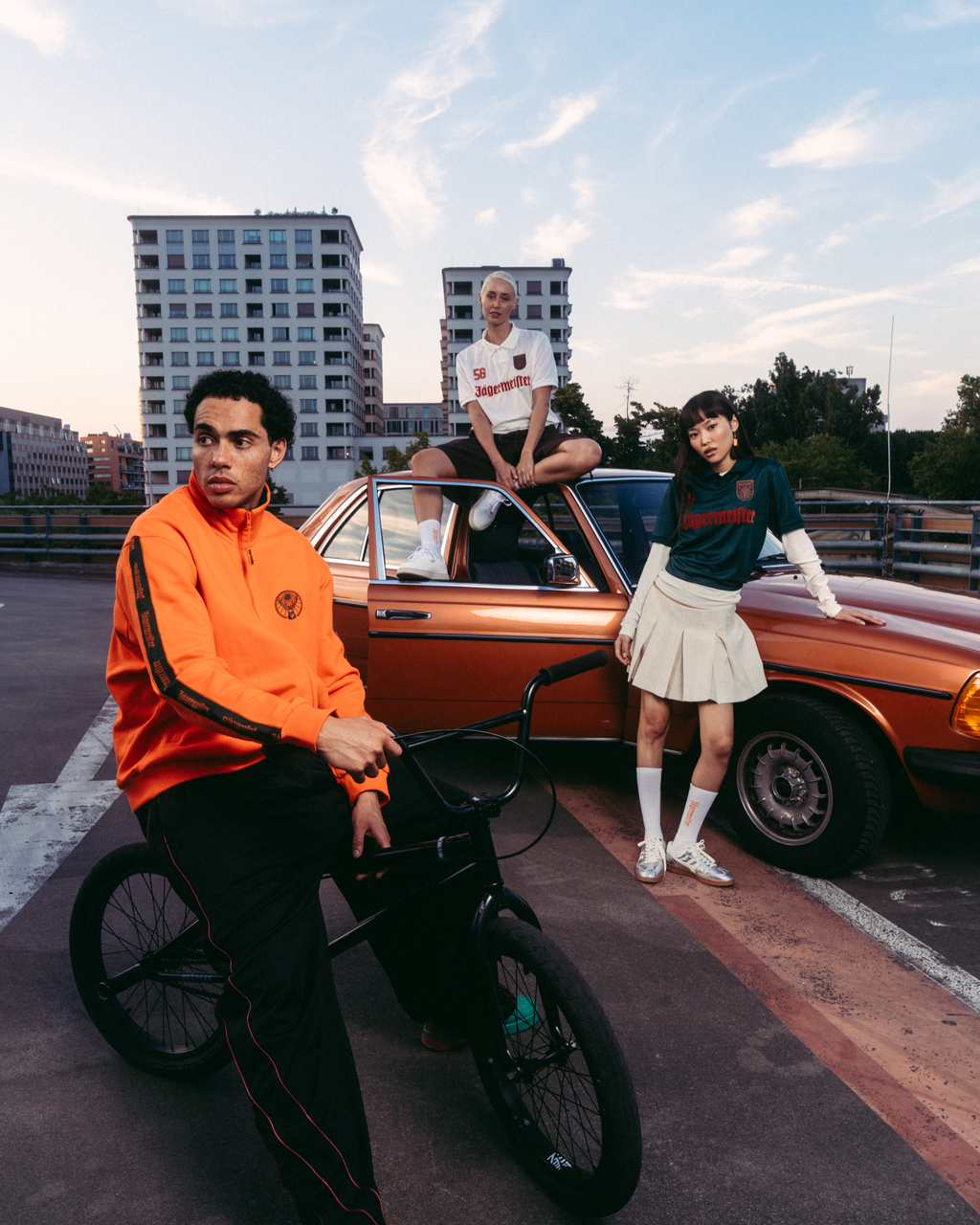 Drie modellen in Jägermeister-streetwear poseren met een vintage oranje auto en een zwarte BMX-fiets op een stedelijk dak bij zonsondergang. De outfits omvatten een feloranje Jägermeister-zipperjack, een zwarte trainingsbroek met rode strepen, een wit Jägermeister-shirt, een groen Jägermeister-shirt met oranje branding en een witte geplooide rok.