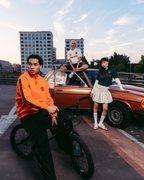 Drie modellen in Jägermeister-streetwear poseren met een vintage oranje auto en een zwarte BMX-fiets op een stedelijk dak bij zonsondergang. De outfits omvatten een feloranje Jägermeister-zipperjack, een zwarte trainingsbroek met rode strepen, een wit Jägermeister-shirt, een groen Jägermeister-shirt met oranje branding en een witte geplooide rok.