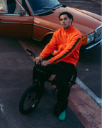 Een groep van vier mensen in een stedelijke buitensetting met een roestige oranje auto, moderne hoogbouw en een brug op de achtergrond. Eén persoon zit op de voorgrond op een fiets in een feloranje Jägermeister-zipper.