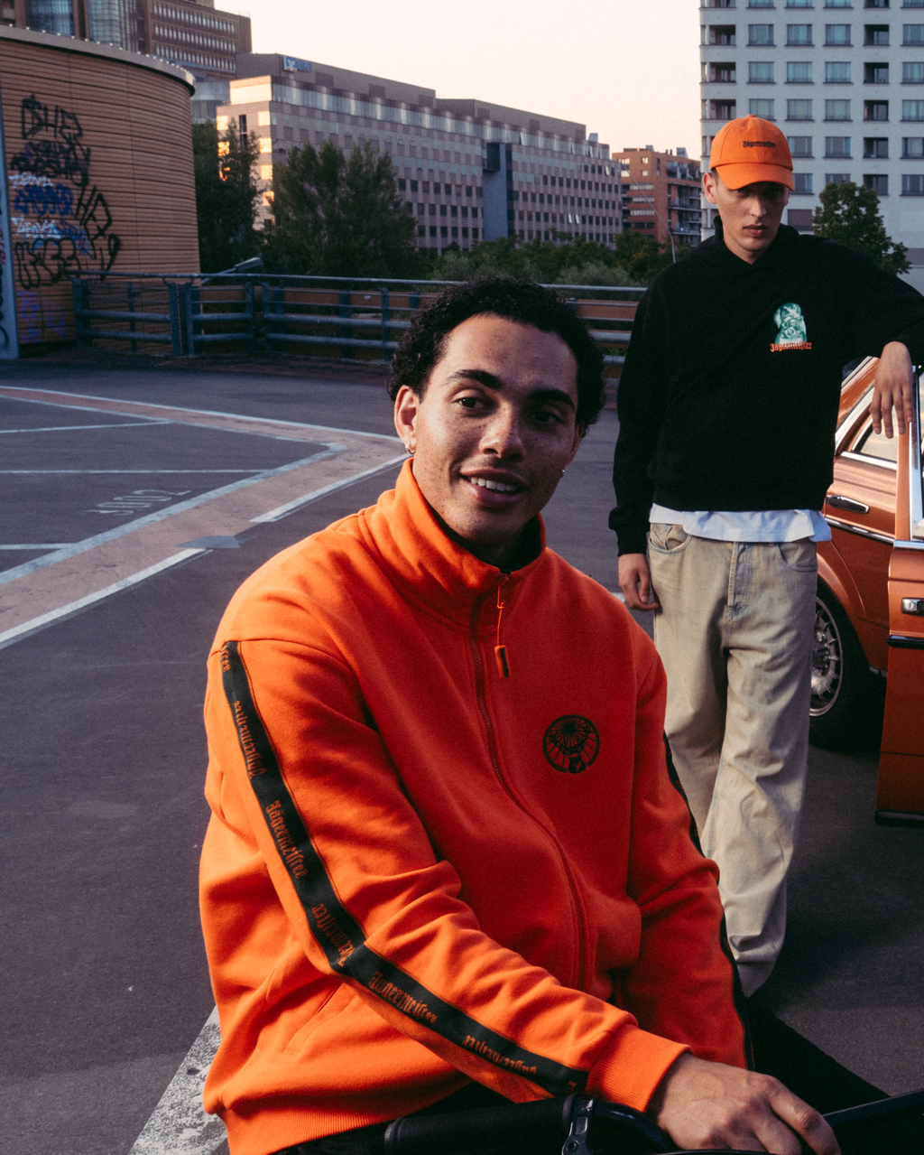 Vier modellen poseren in een stedelijke rooftopsetting met een vintage oranje auto, gekleed in verschillende Jägermeister-streetwearoutfits, waaronder een feloranje Jägermeister-zipper, een wit Jägermeister-T-shirt met Waldi, een donkergroen Jägermeister-shirt en een witte geplooide rok.
