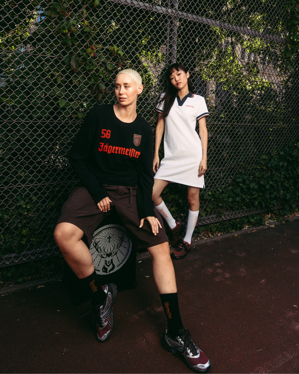 Twee modellen poseren voor een hek van kippengaas. Het model links draagt een zwarte Jägermeister-longsleeve, bruine shorts en zwarte sokken.