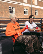 Vier mensen zitten rond een klein tafeltje op een dakterras of brug in de buitenlucht en houden rode bekers vast. Ze dragen casual Jägermeister-streetwear, waaronder een oranje longsleeve en een witte polo.
