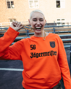 Een lachende vrouw op de voorgrond draagt een feloranje Jägermeister-sweatshirt met lange mouwen en '56 Jägermeister' in zwart op de voorkant, en houdt een gouden Jägermeister-ketting vast. 