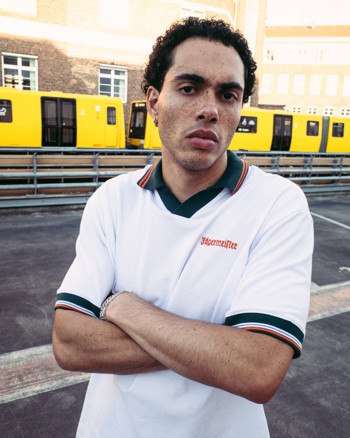 A man wearing a white Jägermeister short-sleeve polo shirt with dark green and orange trim, featuring the brand name 'Jägermeister' on the chest, standing with arms crossed in front of yellow trains and a brick building.