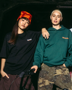 Twee mensen in Jägermeister-kleding poseren binnen tegen een donkere achtergrond. De ene persoon draagt een zwarte oversized T-shirt met een klein Jägermeister-logo en een rood-oranje beanie met patroon, terwijl de andere een groene Jägermeister hoodie draagt met een camouflagebroek. Op de voorgrond is een fietsstuur zichtbaar en de scène is verlicht met gerichte, sfeervolle belichting die de kleding en branding benadrukt.