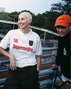 Vier mensen staan buiten op een brug of loopbrug in Jägermeister-streetwear. Eén persoon draagt een feloranje jas met rits, een ander draagt een donkergroen Jagermeister-shirt met een witte rok, een derde draagt een wit Jagermeister-shirt met donkere shorts, en een vierde draagt een zwarte Jägermeister hoodie en een oranje pet.