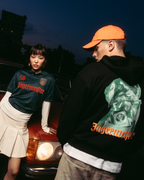 Twee modellen poseren voor een vintage rode auto in de schemering. Eén model draagt een donkergroen 'Jägermeister'-shirt met een witte geplooide rok, en de ander draagt een zwarte Jägermeister Waldi hoodie en een oranje pet.