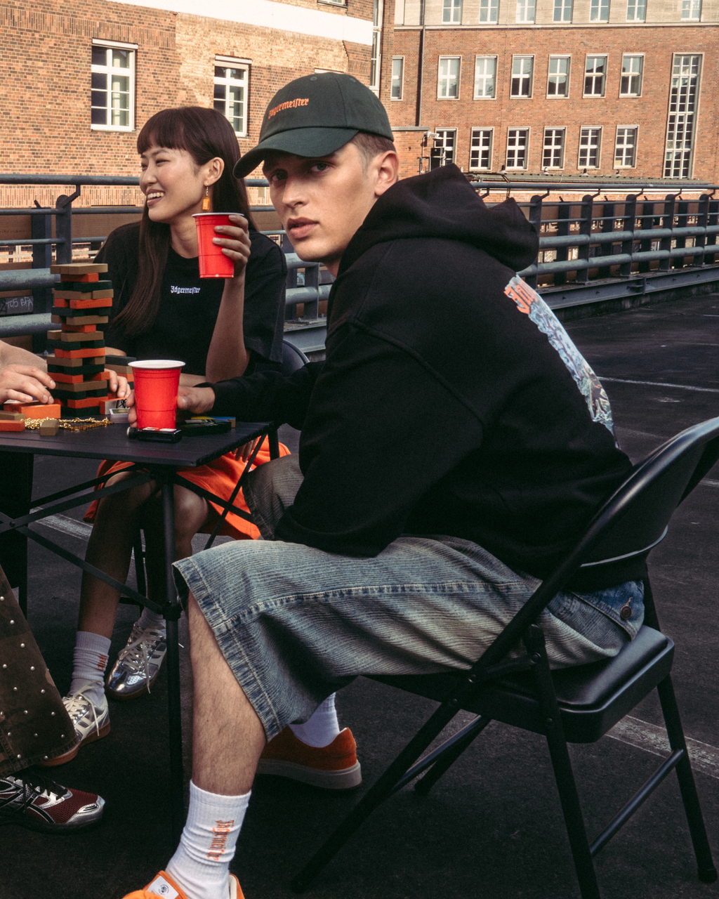Vier mensen zitten rond een kleine tafel op een buitenroof of brug en houden rode bekers vast. Ze dragen casual Jägermeister-streetwear, waaronder een oranje sweatshirt, een wit T-shirt, een zwarte hoodie en een broek met patroon.