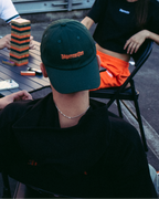 Een glimlachende vrouw op de voorgrond draagt een feloranje sweatshirt met lange mouwen met '56 Jägermeister' in het zwart op de voorkant en houdt een gouden ketting vast. Op de achtergrond zitten drie mensen aan een tafel op een dakterras of buitendek, met een bakstenen gebouw erachter.