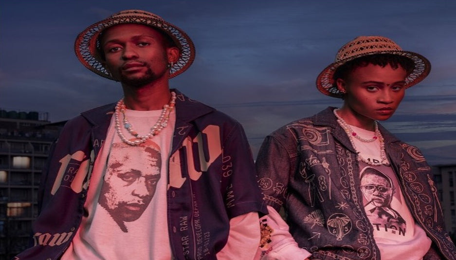 Two models wearing matching graphic jackets, graphic t-shirts, and straw hats with gold jewelry, standing against a twilight sky background.