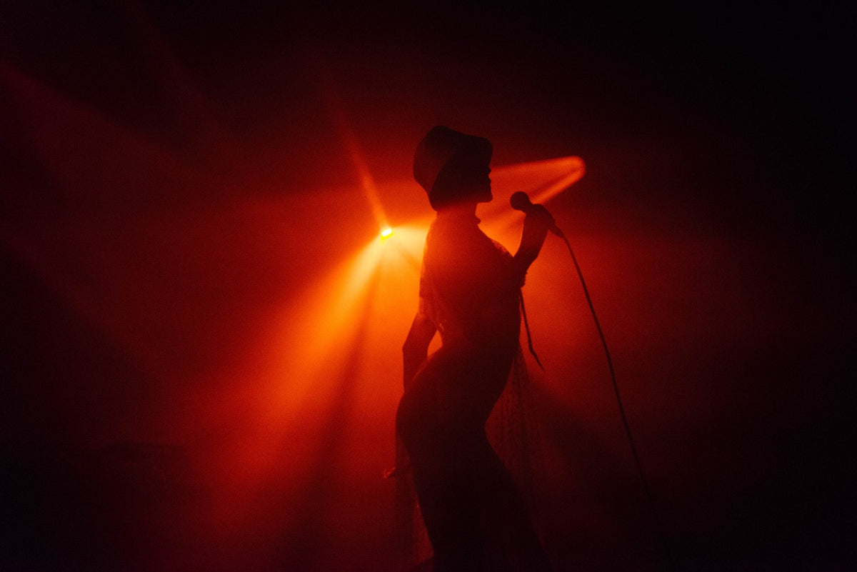 Silhouette of a person holding a microphone on stage, illuminated by dramatic red and orange spotlights in a dark performance setting.