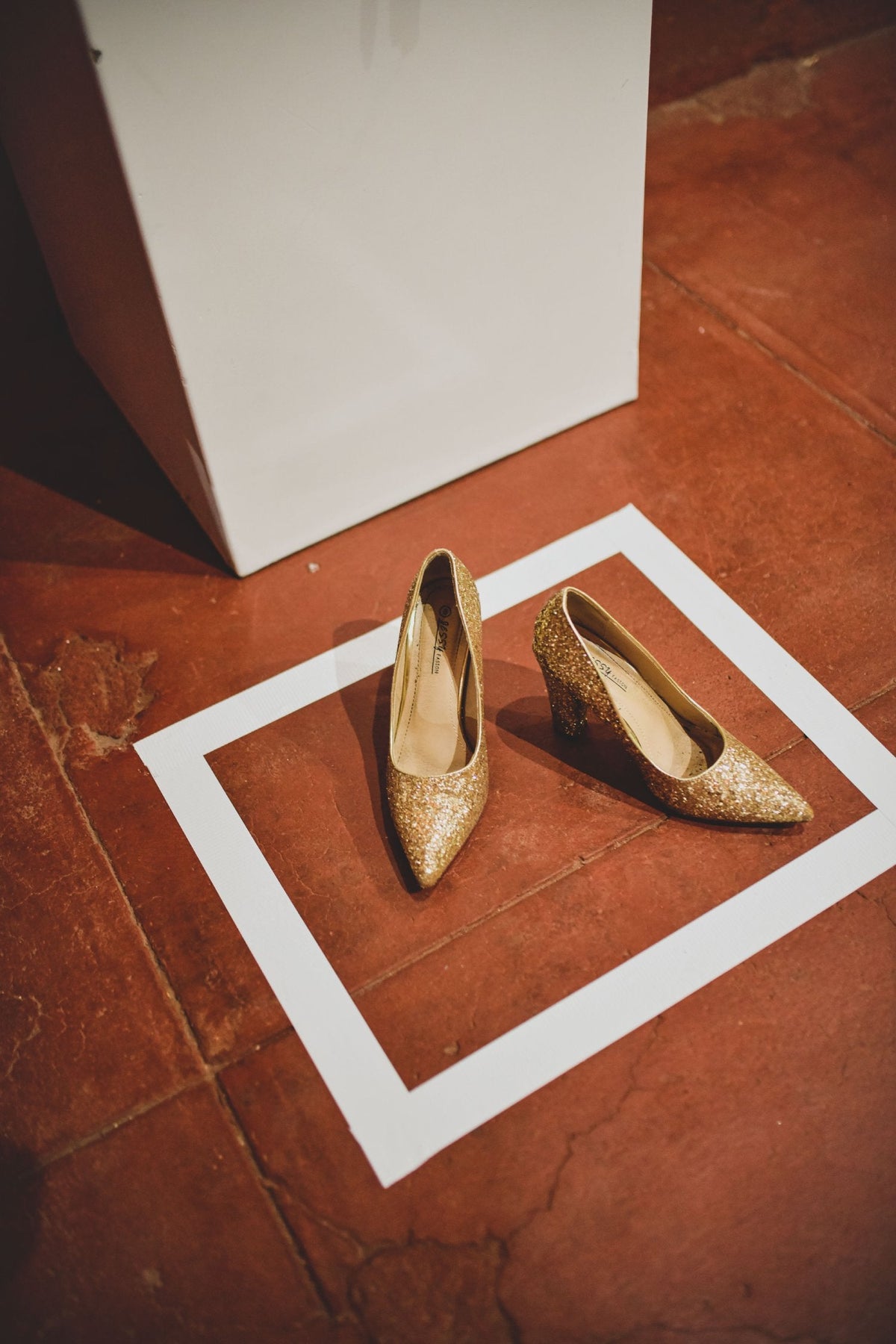 Pair of glittering gold high-heeled shoes arranged on a terracotta tile floor within a white square frame, photographed in warm indoor lighting.