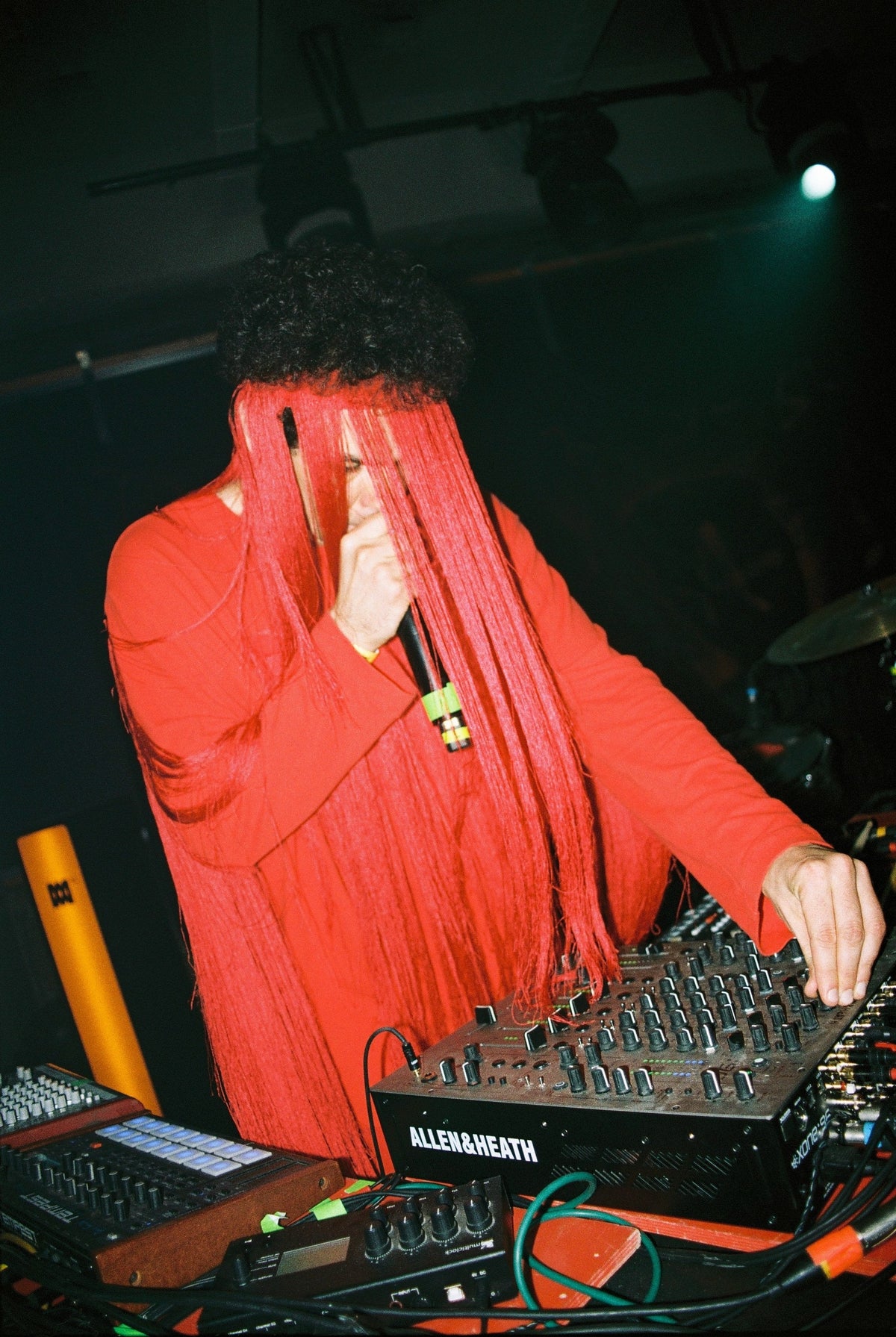 A person in a bright red long-sleeve top and matching red hair/face covering stands behind electronic music equipment, including a mixing board labeled ALEXA HEATH, in a dark stage environment.