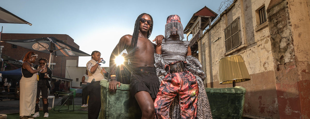 A shirtless man with long braids and a woman in ornate patterned clothing pose together outdoors in front of a building at sunset, with other people and street-side elements visible in the background.