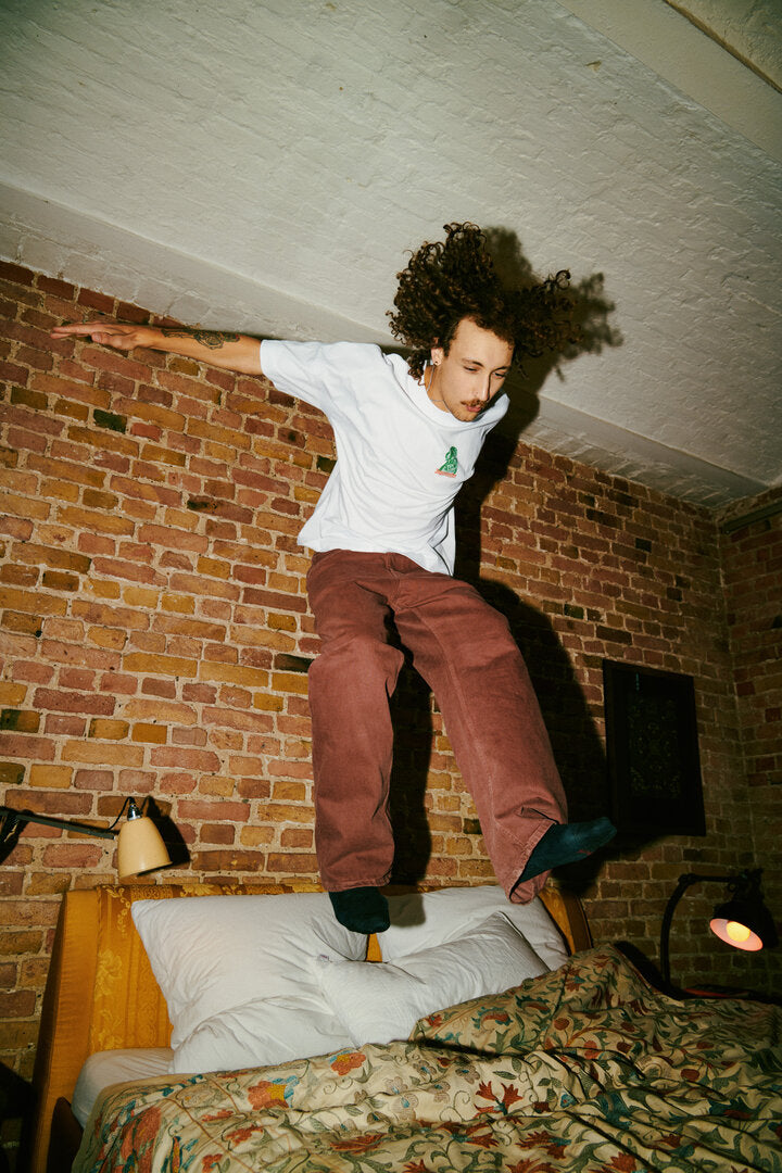 A person jumps onto a bed in a bedroom with exposed brick walls, wearing the Jägermeister Waldi T-shirt and loose brown pants.