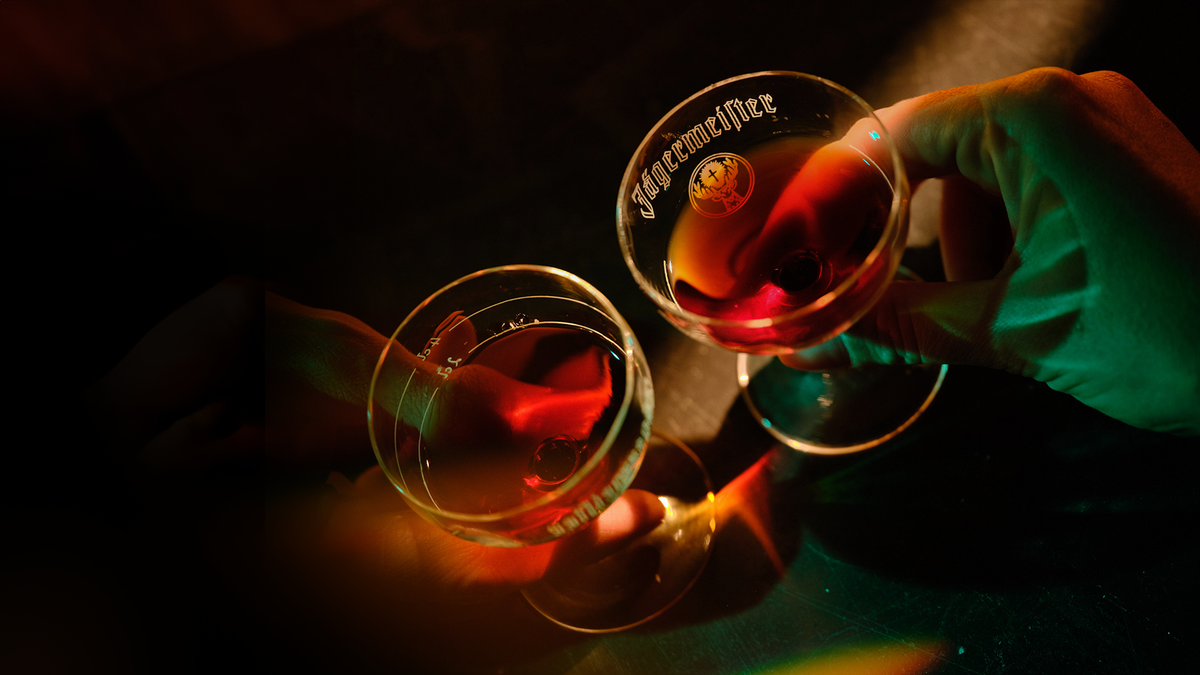 Two stemmed glasses clinking together in a dimly lit setting, filled with a red-orange liquid. One glass shows the word 'Jägermeister' printed on it, and a hand is holding the right glass.